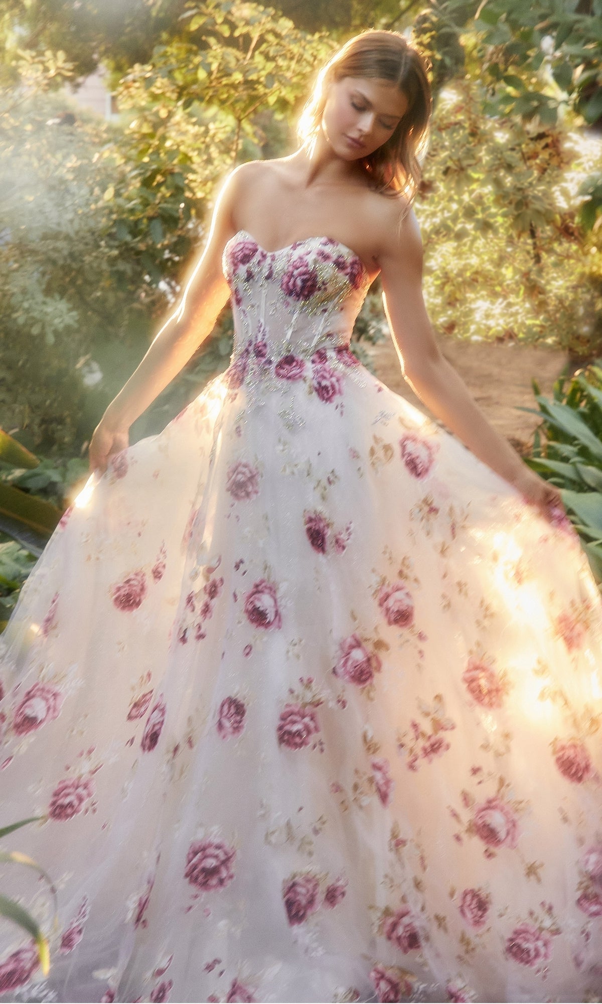 A woman stands outdoors in sunlight wearing the Puff-Sleeve Strapless Floral-Print Ball Gown A1133, gracefully holding out the skirt of her dress.
